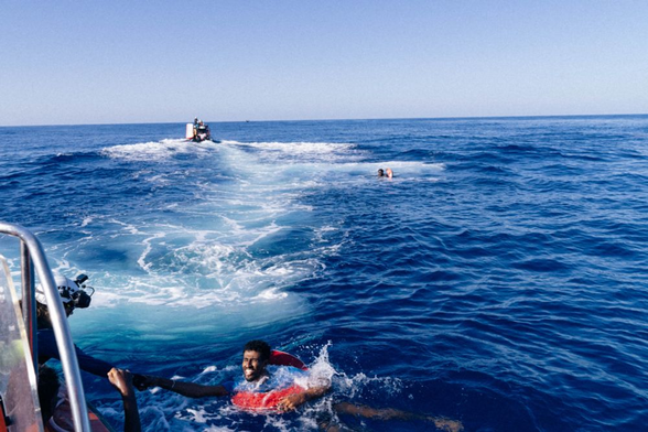 Mitten auf dem Meer. Ganz vorne im Bild reicht eine Person mit Schutzhelm bon einem Boot aus einer mit einem Rettungsring im Wasser treibenden Person die Hand. Ein Stück weit dahinter im Zentrum des Bildes treibt eine weitere Person, die sich mit einem Arm in einen Rettungsring eingehängt hat. Noch weiter dahinter ist ein RHIB von hinten in zügiger Fahrt zu sehen. Es bewegt sich auf einen Punkt zu, wo eine weitere Person oder Gegenstand treibt. Etwas versetzt dazu scheint noch eine Person oder Gegenstand zu treiben. Am Horizont dahinter ist ein größeres Boot / kleines Schiff schemenhaft auszumachen.