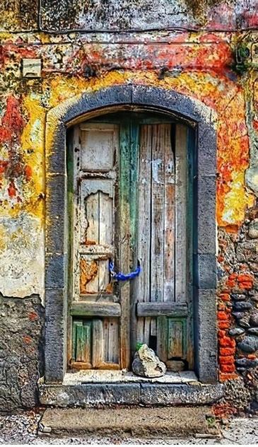 An old wood door on an old building. The door itself doesn't look like it could keep anyone out on account of the dry rot but it is chained shut regardless. The building is made of stone and the painted plaster is falling away exposing layers of paint that resemble fire