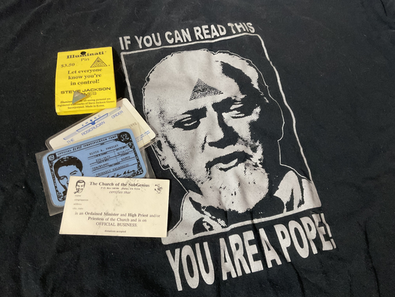 Black t-shirt with the text "If you can read this wyou are a pope!" around Robert Anton Wilson's head, with an illuminati eye in the triangle at his forehead. There's also a Steve Jackson Illuminati enameled pin, an AMORC membership card (expired), an Official Elvis Identification Card (from the 24hr Church of Elvis, PDX), and a The Church of the SubGenius ordained minister and high priest card.