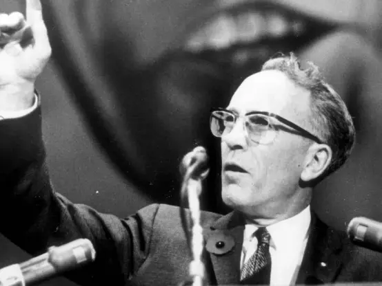 Black and white photo of Tommy Douglas speaking at a podium. Douglas, a key figure in Canadian social democracy and the father of Medicare, is seen with his hand raised.