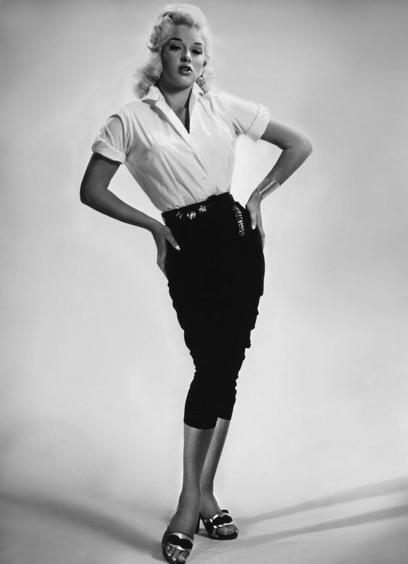 Diana Dors poses confidently, wearing a white shirt and a black pencil skirt. Her hair is styled in waves, and she accessorizes with earrings and bracelets. The image is in black and white, highlighting a classic fashion aesthetic.