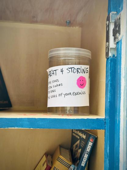 Inside shelf of a Little Free Library in my neighborhood. A few books are visible, as is the clear plastic storage canister I placed next to them. A sign taped to the canister reads: “GREAT 4 STORING”

• Bath Salts 
• Cotton Swabs
• Tea Bags
• The souls of your enemies