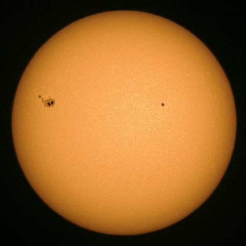 A bright sun with a uniform orange surface, featuring a large sunspot region visible on the left side and a smaller just off from center.