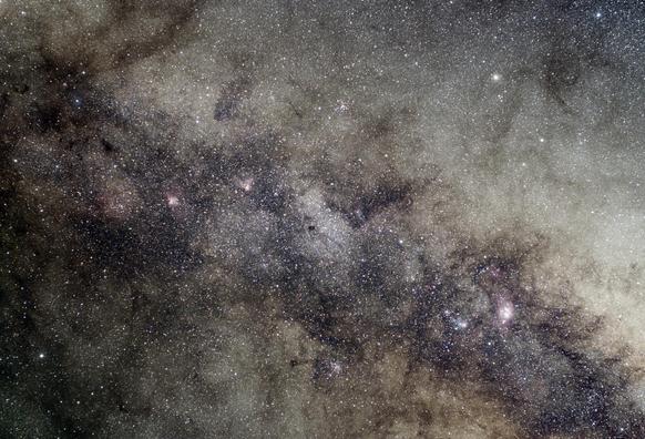 Wide view of the core of the milky way. A band of tiny stars runs from top left to bottom right, and is overlaid with dark dust clouds. In the middle, a hole in the dust, shows the stars beyond. Well known nebulae sit as small-ish blobs in the dust either side of this hole. The swan and eagle on the left, the trifid and lagoon on the right.