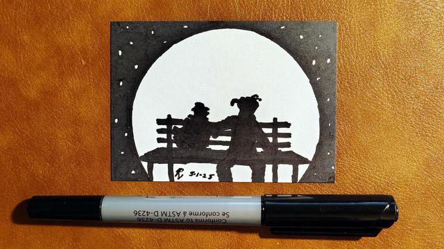 An Artist Trading Card illustrated with a marker drawing of Gonzo and Camilla sharing a tender moment on a bench, shown in silhouette against the moon and a starry sky. The marker used to draw it is laid on the table below the card. The card is signed with my "RV" signature glyph and dated 5-1-25.