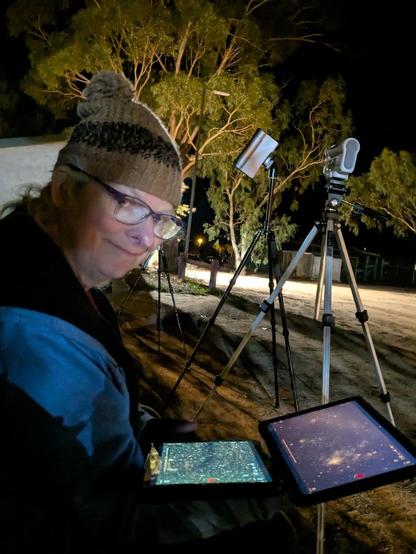 In the foreground a woman is sitting on the red gravelly ground of a caravan park Her head is cocked up at the camera and she is smiling with her mouth closed. Her hair is unkempt, blond and contained by a woolly beanie which is brown striped with a pompom. She has a blue coat on, glasses and nursing two Android tablets controlling her telescopes. One has the Tarantula nebula showing, the other Sandqvist 149. In the mid ground two Dwarf Smart telescopes can be seen on tripods. The closest is set straight on the tripod and is a Dwarfii, the furthest is set in Equitorial Mode and is a Dwarfiii. In the back ground large eucalyptus trees are illuminated.