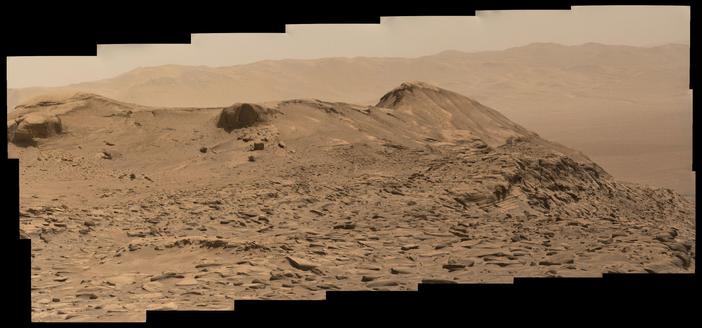 An ochre-toned mosaic capturing a butte located on the slopes of Mount Sharp on Mars, with the wall of the rim of Gale crater in the background.