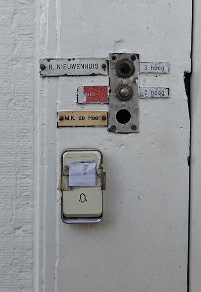 A ln old broken metal doorbell panel with separate name plates as well as plates for the upper floors (3 hoog, 2 hoog). The panel is mounted in a white door frame and has three buttons. A newer plastic doorbell button with a bell icon has been placed a bit below.