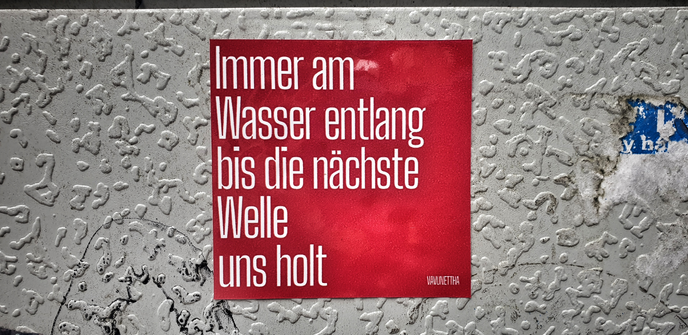 Foto mit HDR-Effekt. 
Die weiße Seite eines Stromkastens.
Darauf ein roter quadratischer Aufkleber mit weißer Schrift:

Immer am
Wasser entlang
bis die nächste
Welle
und holt


Unten rechts kleiner: Vavunettha