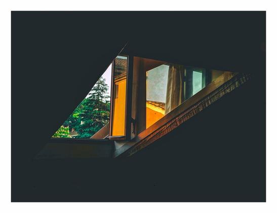 A photo of a window and mirror at sunset. They are positioned it such a way that they appear to be two triangles stuck together creating a rhombus like shape. Outside the window you can see some green trees and a bit of sky. The mirror reflects some yellow buildings. The rest of the photo is black and highlights the weird shape of the objects.