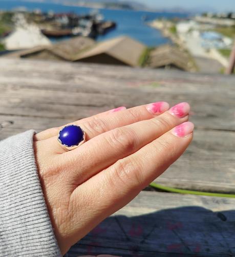 a silver open ring with a big blue stone, it's worn on a hand with pink finger nails