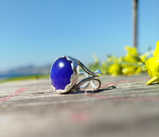 a silver open ring with a big blue stone, placed on wooden floor with yellow flowers on background left