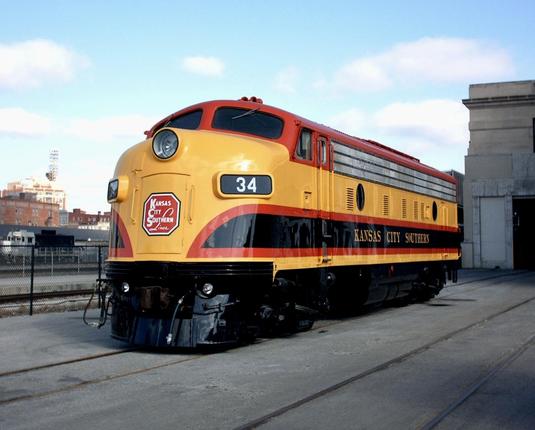 An old and sleek looking locomotive engine in yellow red and black colors. On the front is the Kansas City Southern stop sign logo