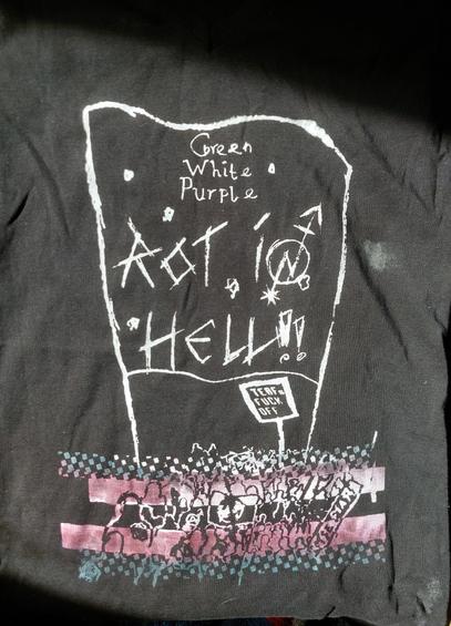 The motive, printed on black textile, consists of a crude drawing of a banner, featuring the text 'Green White Purple, Rot In Hell', and a small sign underneat it which reads 'TERFs Fuck Off'. Banner and sign are held by people who are part of a big, not very detailed crowd of people, printed in the colours of the Trans* Pride Flag.