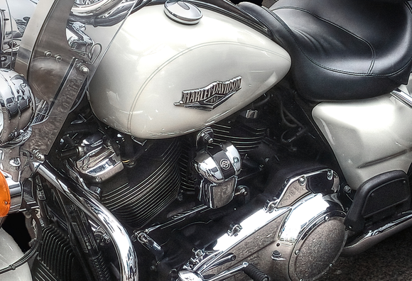 A wall art piece, an image of a closer look at part of an iconic white Harley Davidson Motor cycle, featuring the signature badge and shining chrome shapes, a black leather curved seat and black engine parts.