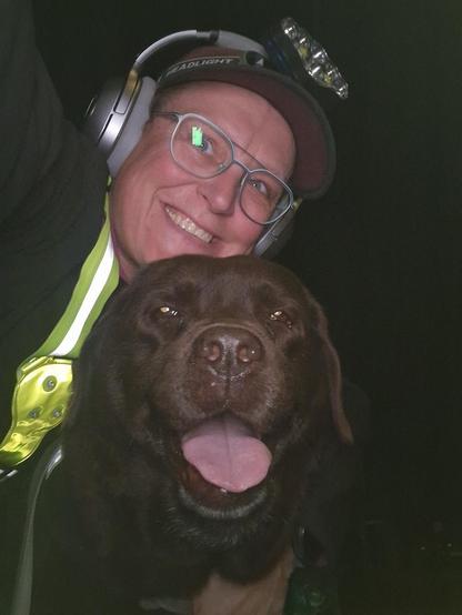 Night time selfie of a person and a chocolate brown Labrador with greying snout. The person is wearing a black jacket, yellow safety harness, red cap, silver headphones and blue-rimmed glasses. On their cap rests a headstrap torch in off position. Their free hand is around the dog's shoulders. Both are facing the camera. The dog's mouth is slightly open, showing off some blep. The person's head is resting on the dog's head. They're smiling, grateful for their time together.