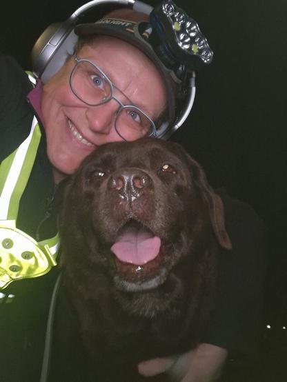 Night time selfie of a person and a chocolate brown Labrador with greying snout. The person is wearing a black jacket, yellow safety harness, red cap, silver headphones and blue-rimmed glasses. On their cap rests a headstrap torch in off position. Their free hand is around the dog's shoulders. Both are facing the camera. The dog's mouth is slightly open, showing off some blep. The person's head is resting on the dog's head,. They're smiling, grateful for their time spent together.