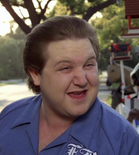 A leering face being made by Pee Wee Herman's nemesis & bike coveter, Francis! Francis is chubby with a large head, pale cream skin, straight brown hair combed back in a poofy helmet. His mouth is open and his tongue is partially out. He looks like he's mocking, which he probably is. This still is a scene from the movie. He's probably saying, "I know you are but what am I!?" (or something). He's wearing a blue camp shirt with gothic script initials, cut off in photo, but they read FB.