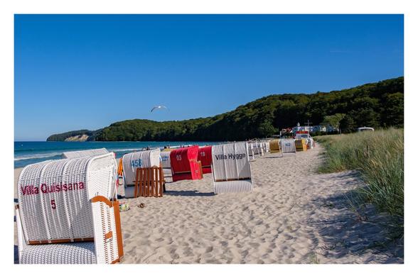 Foto im Querformat. Blick über einen Strand. Rechts ist Dünengras, mittig der Strand. Weiße und rote Strandkörbe stehen mit dem Rücken zur Kamera. Auf dem vordersten steht Villa Quisisana. Links sieht man das blaue Meer. Am Horizont ist eine Landzunge. Eine Möwe fliegt über den Strand. Es ist sonnig, der Himmel klar und blau.