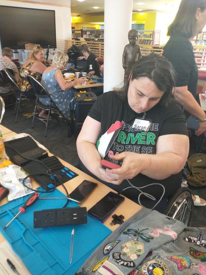Sarah, a woman with light skin tone and long dark hair, sitting in a wheelchair and holding a small electrical which is plugged in. In front of her are the several other phones or small electricals and some screwdrivers. Behind her are other volunteers and attendees working on things at tables.