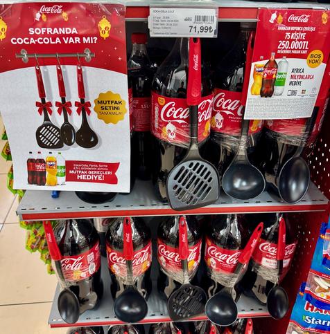 a somewhate perplexing promotional offer of free black plastic kitchen utensils such as serving spoons and spatulas taped to large coke bottles  in a supermarket