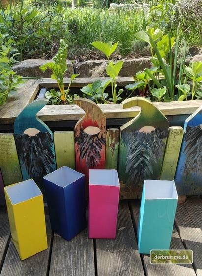 Bunt besprayte Tetrapacks auf einer Holzterrasse. Dahinter Maurerpötte, die von bunt besprayten Holzwichteln umgeben sind.