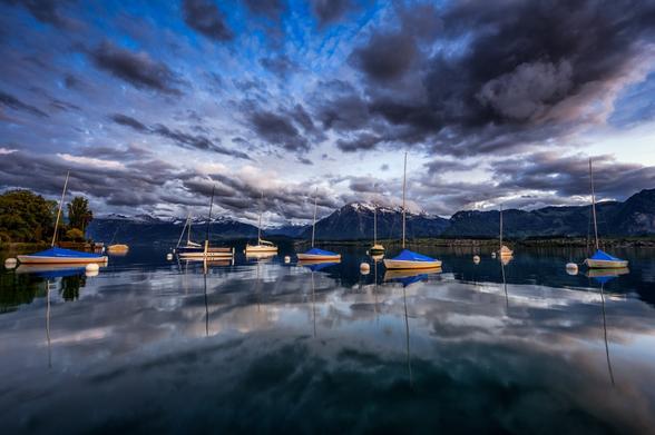 Weitwinkelfotografie von Segelbooten an Bojen auf einem ruhigen See, in dem sich interessantes Wolkenspiel reflektiert.