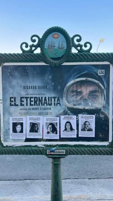 En la foto se ve un cartel enmarcado de la serie de "El Eternauta" en el espacio público. El afiche se compone de un primer plano de un hombre con traje de buzo. Sobre el cartel se ven pegados cinco afiches cada uno con su respectiva foto e información. Son fotos de Héctor Oestherheld y sus cuatro hijas, todos detenidos desaparecidos de la última dictadura en Argentina