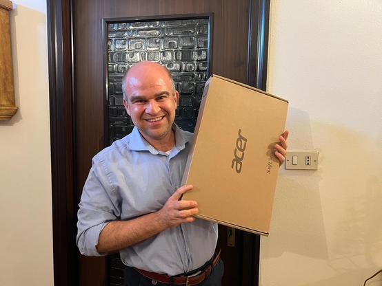 A smiling man in a light blue shirt stands indoors holding a cardboard box with the Acer logo, indicating it contains a new laptop from the Acer Swift Series. He is posing happily in front of a wooden door with decorative glass panels.