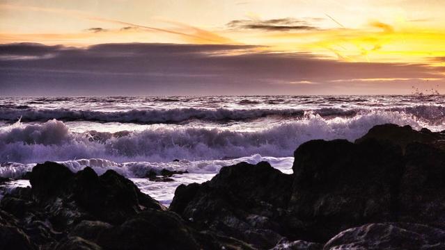 Dramatic image of large white waves crashing against dark, jagged rocks under a vibrant yellow and purple sunset sky.