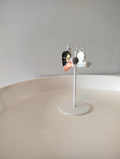 a heart shaped silver pendant with wristwatch elements hanged from a white wire, and a similar pendant which we can see only it's silver back