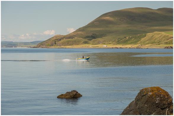 A small boat with a person is navigating across calm waters near a green, hilly shoreline. In the foreground, there are a few rocks visible. The sky is clear with a few clouds.