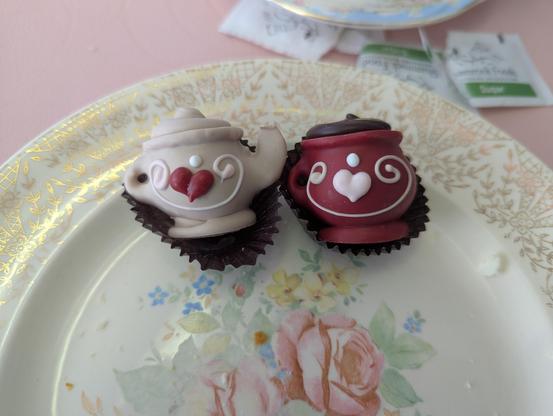 A photo of a plate with two cherry cordials. The left one is a soft grey in color and shaped like a tea pot. It has a decorative design surrounding a red heart. The right one is red and shaped like a mug with a matching design with a pink heart.