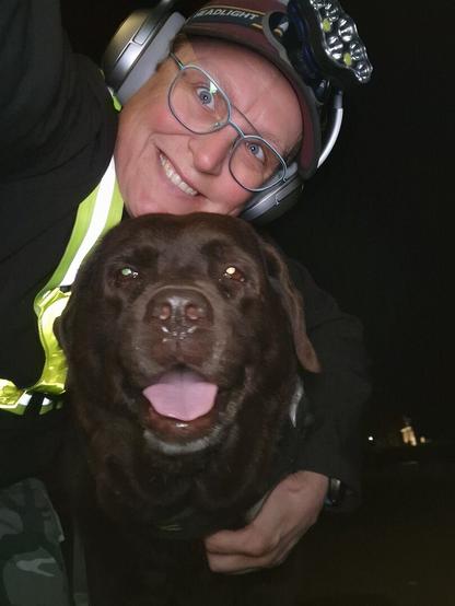 Night time selfie of a person and a chocolate brown Labrador with greying snout. The person is wearing a black jacket, yellow safety harness, red cap, silver headphones and blue-rimmed glasses. On their cap rests a headstrap torch in off position. Their free hand is around the dog's shoulders. Both are facing the camera. The dog's mouth is slightly open, showing off some blep. The person's head is resting on the dog's head. They're smiling, grateful for their time spent together.