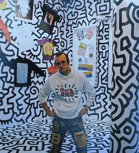 Photograph of Keith Haring standing in the Pop Shop in 1986. Keith is wearing jeans and a white hoodie with his 'radiant baby' printed on it. (A figure resembling a crawling infant with 'beams' around  it.) The Pop Shop has white walls covered in black outlines of Haring's designs. In the background some t-shirts pinned to the wall are visible.