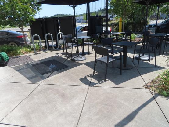 Towards the back are cars going through the drive-thru at Starbucks in Cloverdale, CA. A thin strip of landscaping separates the drive-thru from what's meant to be a 7 bike wave rack. But almost immediately in front of the empty wave style cycle rack is a patio table with chairs that have arms. And there are three other sets of this patio furniture. The photo is taken from the curbcut that leads from the disabled parking.