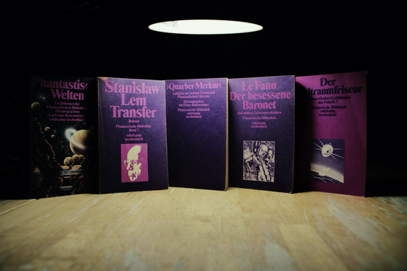Five vintage science fiction and fantasy paperback books from the Suhrkamp “Phantastische Bibliothek” series stand upright in a row on a wooden surface under a circular overhead lamp. The books feature dark purple covers with pink and white text, and include titles by Stanisław Lem and Le Fanu, as well as themed anthologies and essays on speculative literature.