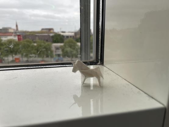 Someone has placed a small origami unicorn on a white windowsill. In the background you can see a blurred silhouette of a city