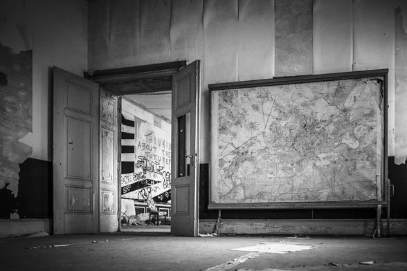 Ein Foto in schwarz-weiß: Blick auf eine große, stehende Landkarte auf der rechten Seite. Links ist eine Tür geöffnet, der Blicken durch zeigt einen weiteren Raum. An der Wand prangt ein Graffiti:” thinking about the future”