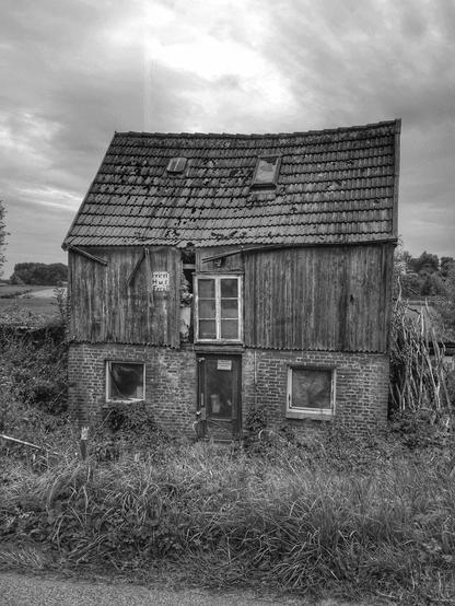 Schwarzweißfoto eines kleinen, verlassenen und verfallenen Hauses am Wegesrand inmitten von hohem Gras. Das Foto zeigt die Fassade mit der Eingangstür im gemauerten Sockel des Hauses. Zu beiden Seiten der Eingangstür befinden sich quadratische Fenster, die von innen verhängt sind. Das obere Stockwerk hat eine hölzerne Fassade, die aus einer vertikalen Bretterverschalung besteht, die an einer Stelle offen und beschädigt ist, links von einer mittig eingebauten Tür. Weiter links von dieser Tür ist ein Schild montiert auf dem geschrieben steht 'Ferien Hus frei'. Oben schließt das Haus mit einem Ziegeldach mit zwei Fensterlucken ab. Eine Dachrinne hängt teilweise herunter. Hinter dem Haus befindet sich ein Feld und Gestrüpp. Der Himmel ist bewölkt.