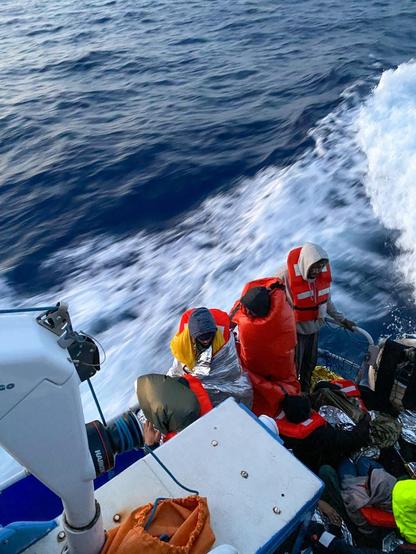 Blick von oben auf die Steuerbordseite des Hecks der AURORA SAR in zügiger Fahrt. Dort stehen an die Reling gelehnt und sitzen auf dem Boden Menschen in, die teilweise zum Schutz gegen Kälte in Schlafsack, Decken oder Rettungsfolien gehüllt sind.