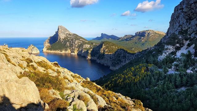 A high-angle shot captures a dramatic coastline with rocky cliffs plunging into a blue sea. Jagged mountains rise in the background under a partly cloudy sky.