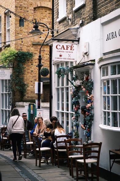 A woman is walking down a lane in Greenwich Market past three tables where three pairs of people are sitting outside a cafe. The nearest couple have a corgi dog sitting on the man's lap. There are fabric flowers and decorations around the cafe door and a waitress is standing in the doorway looking out. Part of the name, "Aura C" can be seen above the cafe window, and a sign above the door reads, "Café Coffee Lunch Ice Cream".