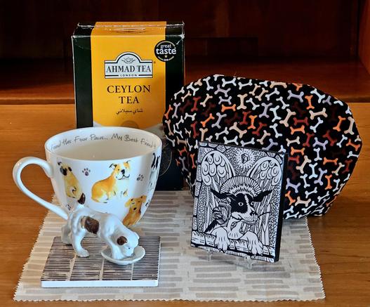 Tea tableau with a dog theme featuring a chewy bone cozy fabric, a tea towel and coaster reminiscent of newsprint, a mug with painted dogs, an ink drawings of a terrier with angel wings, and a vintage porcelain figurine of a dog stepping in its food dish. The tea is Ceylon black loose-leaf from Ahmad.