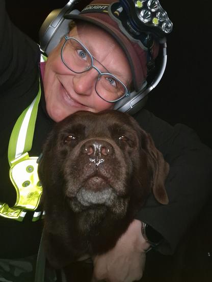 Night time selfie of a person and a chocolate brown Labrador with greying snout. The person is wearing a black jacket, yellow safety harness, red cap, silver headphones and blue-rimmed glasses. On their cap rests a headstrap torch in off position. Their free hand is around the dog's shoulders. Both are facing the camera. The person's head is resting on the dog's head. They're smiling, grateful for their time together.