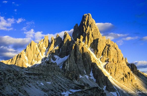 Strong golden side-lighting picks out the complex ridges and gullies of a steep rocky peak with a few streaks of snow against a deep blue sky.