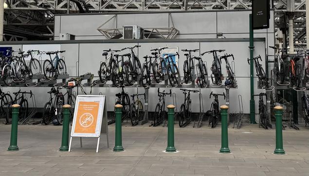 Waverley Station: In the foreground, six green bollards with gold caps are joined by a sandwich board with an orange poster stating ‘No e-bikes or e-scooters to be stored in the station’. Behind, a two-deck cycle store featured a range of parked up cycles, around 29 in shot.