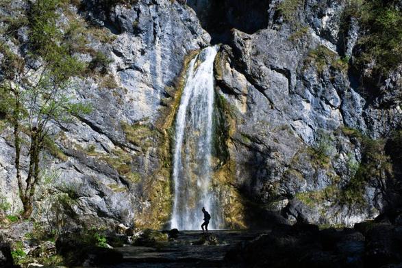 Ein vierzig Meter hoher Wasserfall, der durch eine Felsspalte hervortritt und in ein Wasserbecken abfällt. Vorne geht eine Frau durchs Bild und steht direkt vor dem Wasserfall.