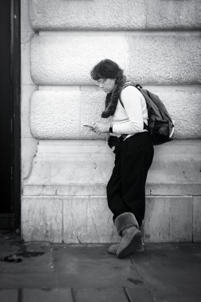 Black and white. A lady tourist in her late forties/early fifties is leaning on a white stone wall, facing to the left, looking at her phone. She has neck length brown hair, wears glasses, and is dressed in a white knitted jumper, a thick furry neck-warmer, black walking trousers, and what look like Ugg boots. She has a bum bag at the front and a backpack. She looks like she's set for walking around London.