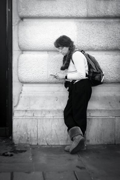 Black and white. A lady tourist in her late forties/early fifties is leaning on a white stone wall, facing to the left, looking at her phone. She has neck length brown hair, wears glasses, and is dressed in a white knitted jumper, a thick furry neck-warmer, black walking trousers, and what look like Ugg boots. She has a bum bag at the front and a backpack. She looks like she's set for walking around London.